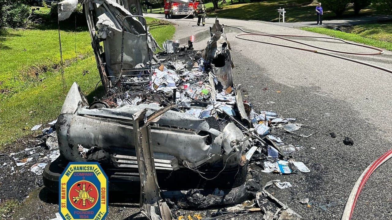 A heavily damaged vehicle burned to debris on the side of a road, with fire hoses nearby and emergency responders visible in the background. The scene indicates a recent fire incident.