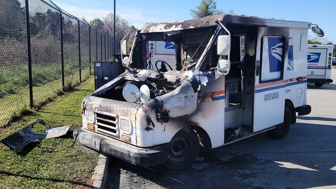 A damaged postal delivery truck with fire damage to the front. The vehicle shows signs of severe burns, with soot and charred metal visible on the exterior. A fenced area is seen in the background.