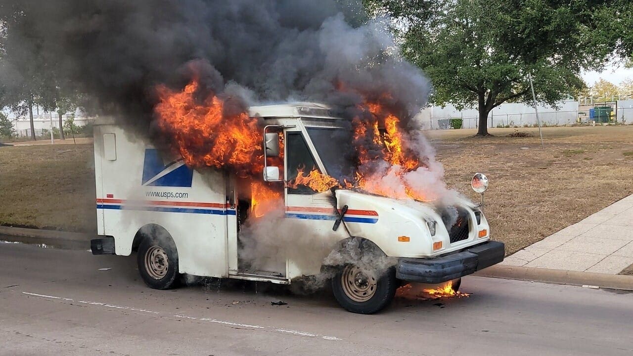 A postal service delivery truck engulfed in flames, producing thick smoke, parked on a street with trees in the background. The fire appears to be intense and the situation looks severe.