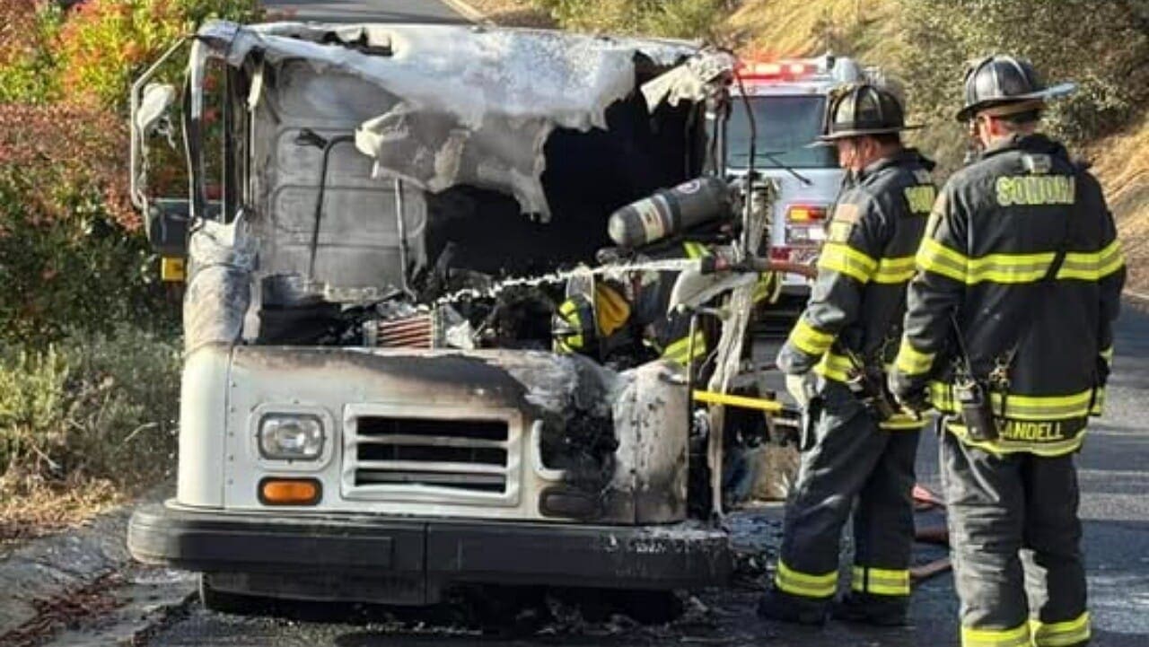 Firefighters inspecting a burnt delivery truck on the side of the road. The vehicle has significant fire damage to the front, and emergency response vehicles are visible in the background.