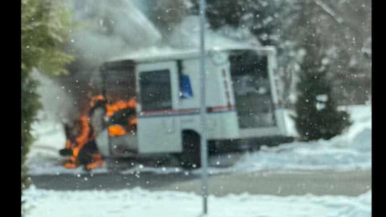 A postal delivery truck is engulfed in flames with smoke rising, parked on a snow-covered street. Nearby trees and a street sign are partially visible.
