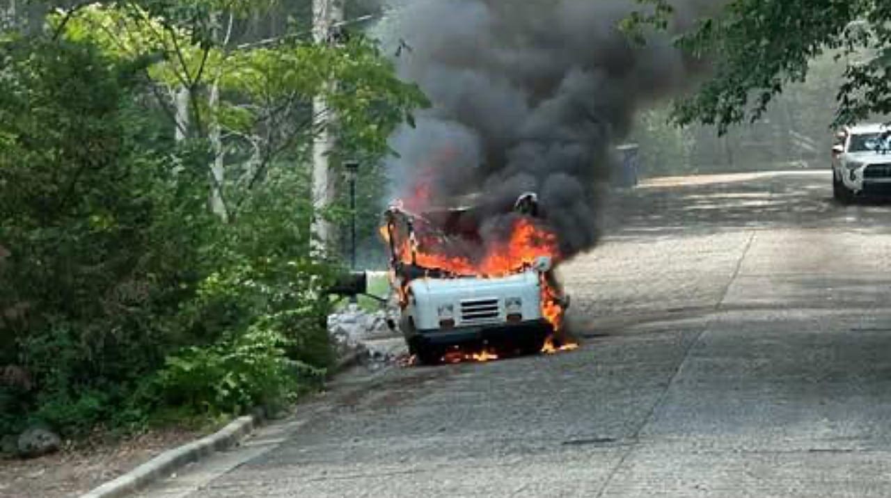 A vehicle on fire on the side of a road, with flames and thick smoke rising into the air. Trees and foliage are visible nearby, along with a second vehicle parked further down the road.