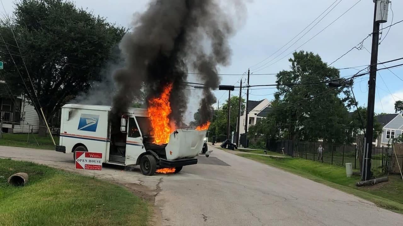 A postal delivery truck with flames and smoke coming from the back, parked on a residential street with nearby trees and houses.