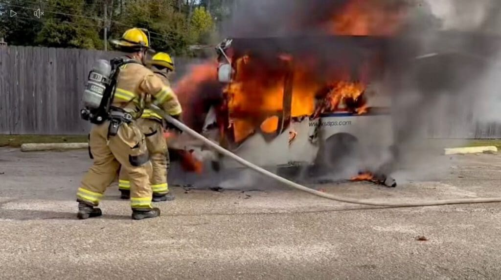 Two firefighters in protective gear using a hose to extinguish a small vehicle fire, with flames and smoke billowing from the vehicle, set against a wooded background.