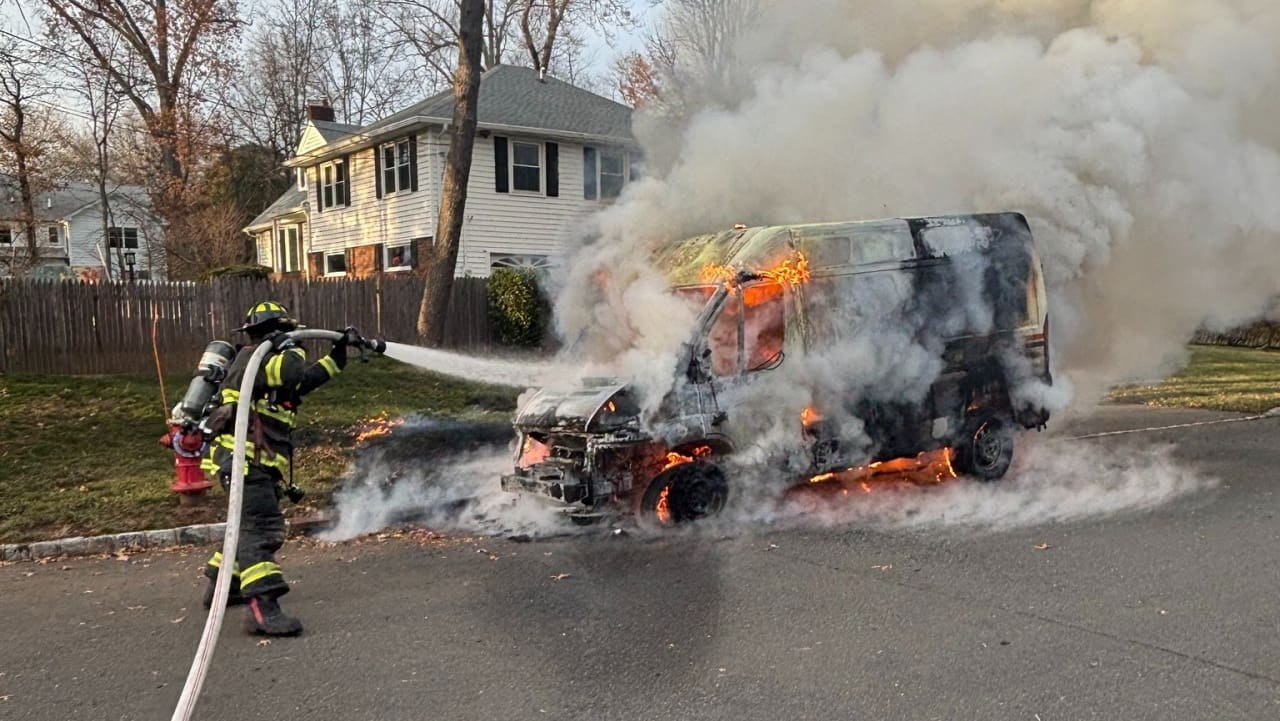 A firefighter spraying water at a van engulfed in flames and thick smoke on a residential street, with houses in the background.