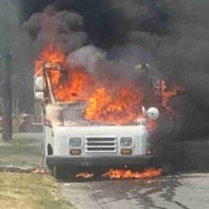 A delivery truck engulfed in flames, producing thick black smoke, parked on the side of a road. The nearby area appears clear of people.