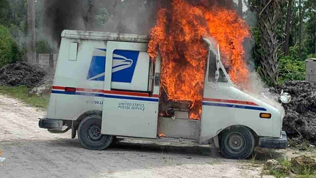 A United States Postal Service delivery van engulfed in flames, with fire visible at the front door. The surrounding area features dirt and some greenery.