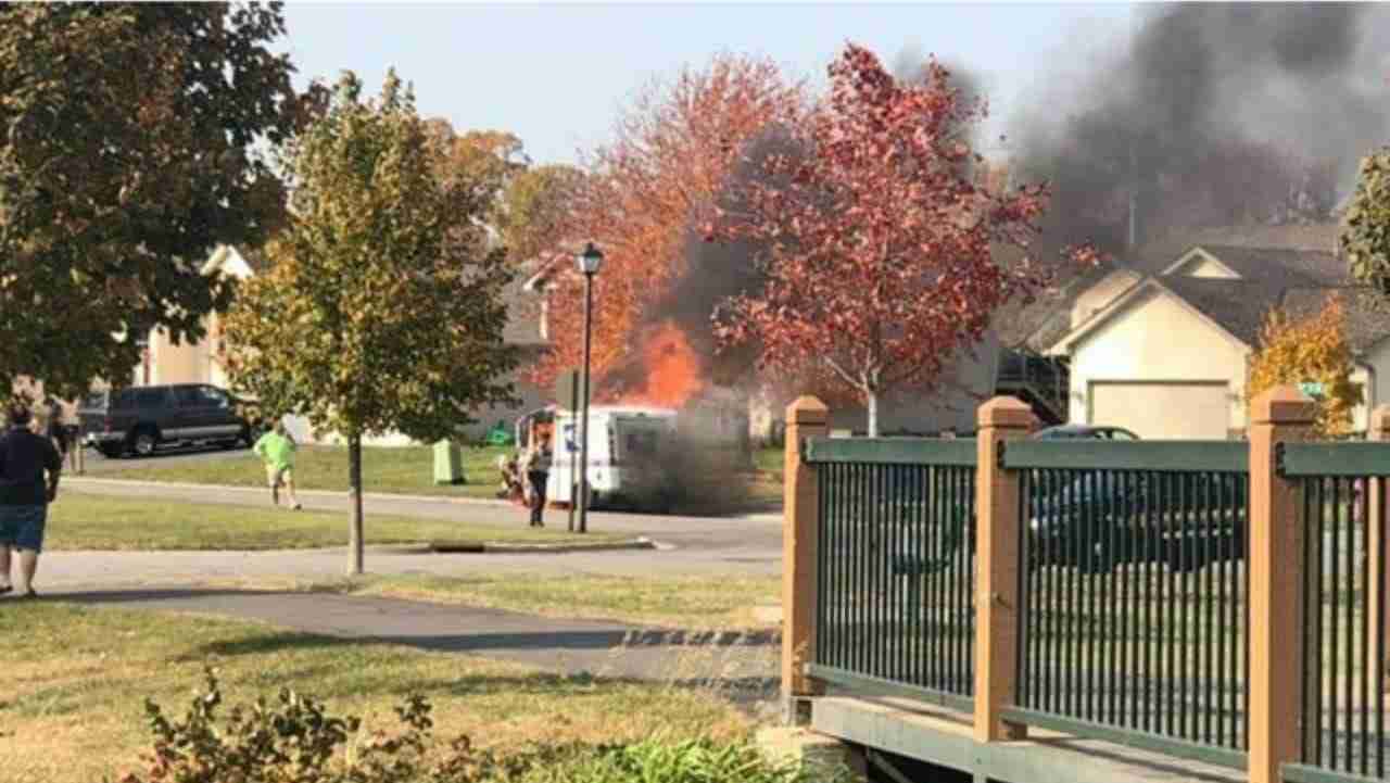 A van is on fire in a residential area, producing thick black smoke. Nearby, people are standing and watching. Trees with autumn foliage are visible in the foreground and background, along with houses in the neighborhood.