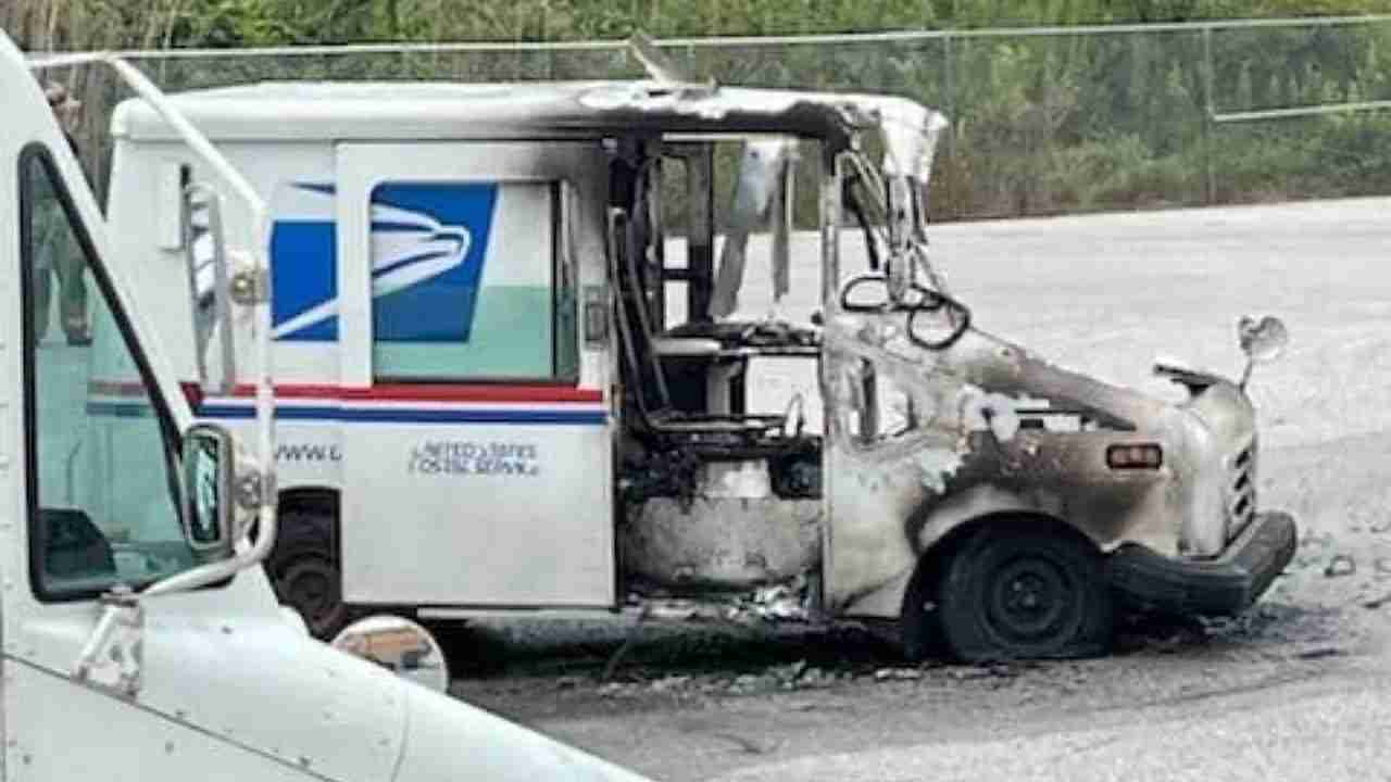 A damaged United States Postal Service delivery truck with a burnt exterior, showing extensive charring and destruction on the front section, parked on an asphalt surface. Another postal truck is partially visible in the foreground.