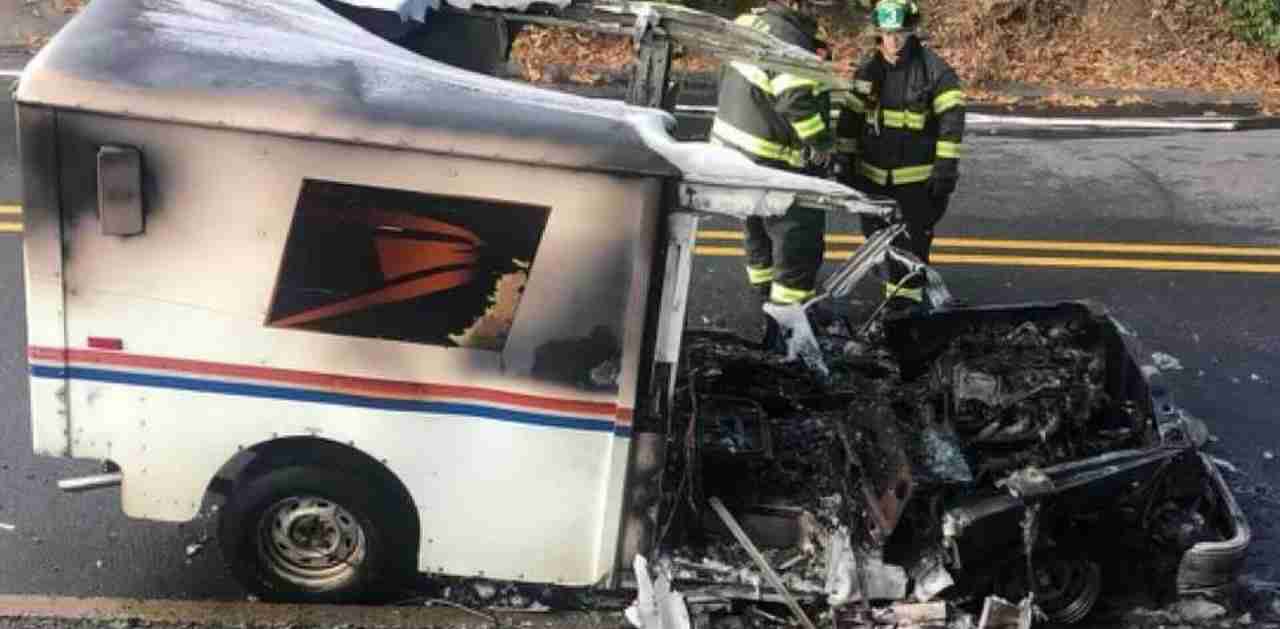 A damaged postal service vehicle with extensive fire damage, showing charred remains over a roadway. Two firefighters in full gear stand next to the vehicle, which has burnt components visible.
