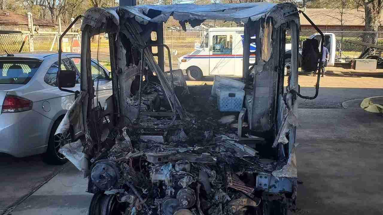 A burned-out delivery vehicle with significant damage to the front and interior, parked next to a white car in a lot. A postal service vehicle is visible in the background.