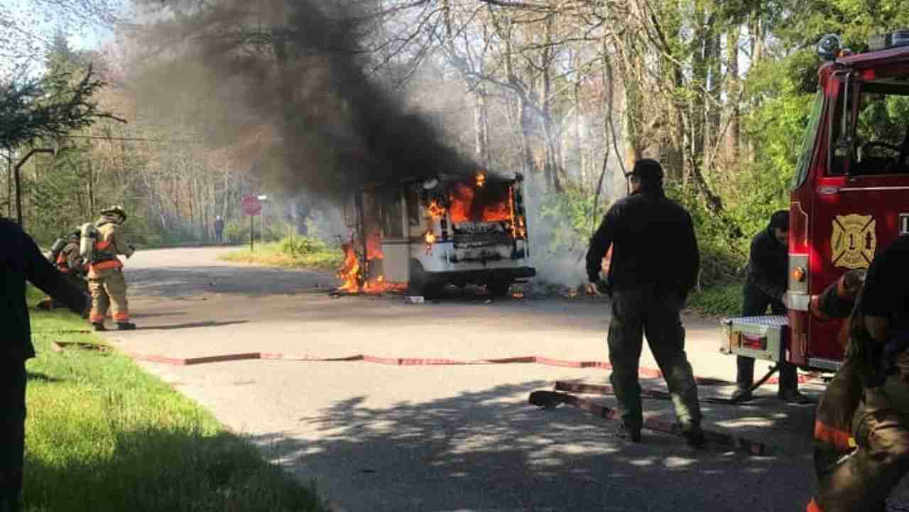 A fire truck parked on the side of the road near a burning vehicle releasing thick black smoke. Firefighters are attending to the fire while onlookers observe from a safe distance.