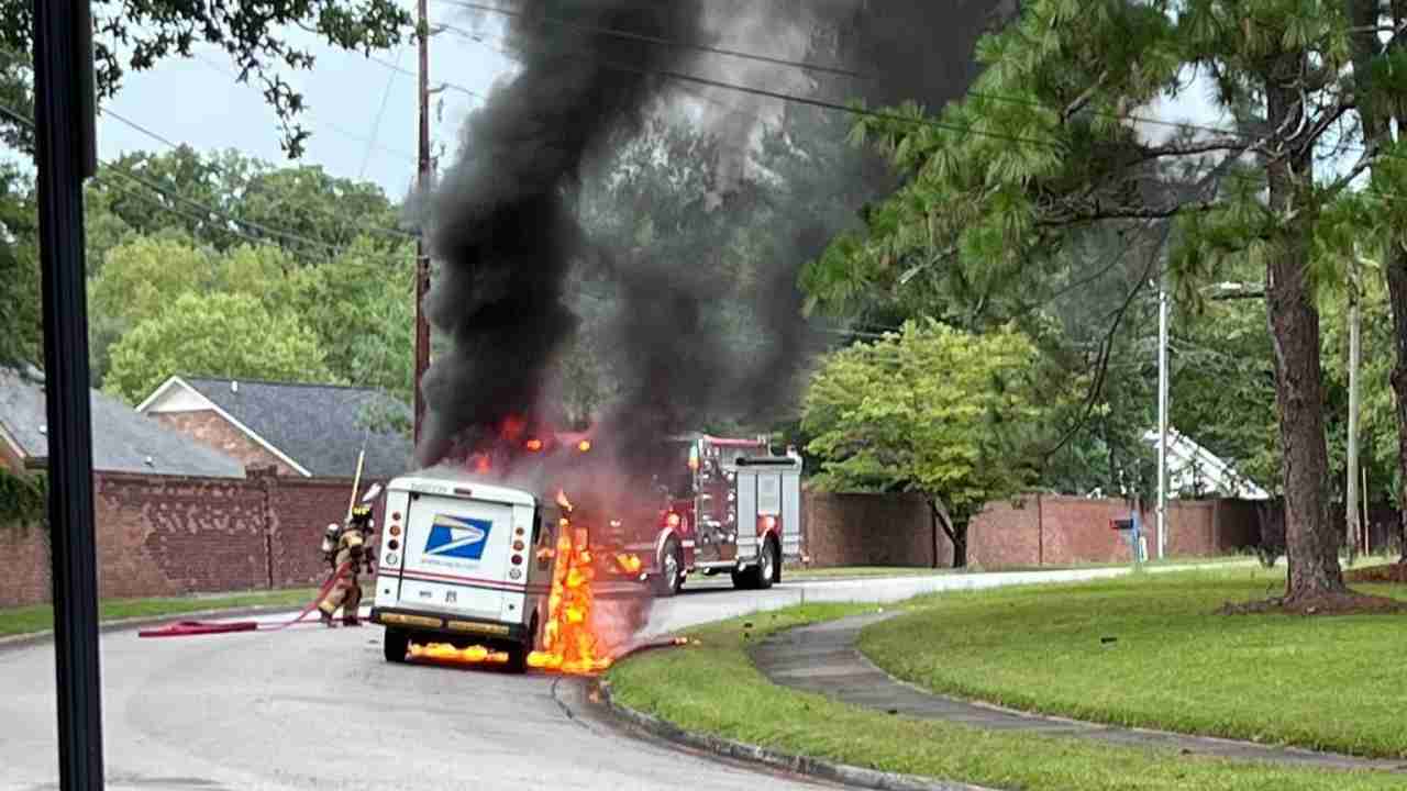 A USPS delivery truck on fire, emitting thick black smoke, on a residential street. Firefighters are present, attempting to control the flames. Green trees and brick walls provide context of the neighborhood.