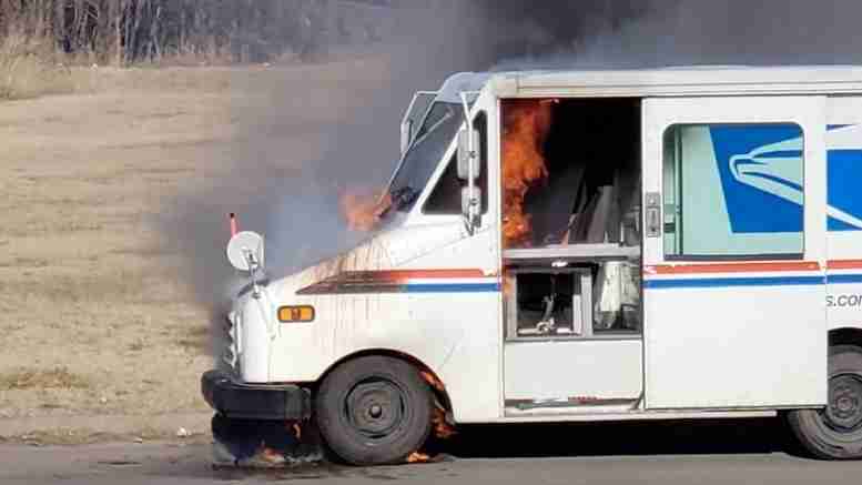 A postal delivery truck with flames and smoke rising from the front, indicating an active fire, on a grassy area.