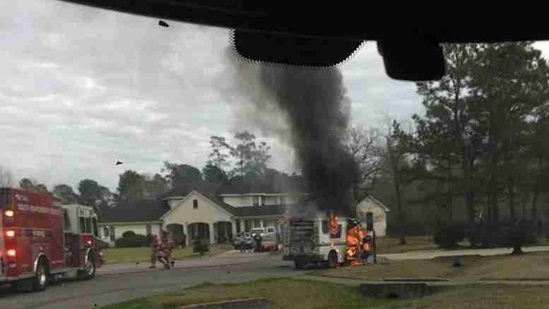 A fire truck and firefighters respond to a vehicle engulfed in flames, emitting thick black smoke, near residential homes on a street.