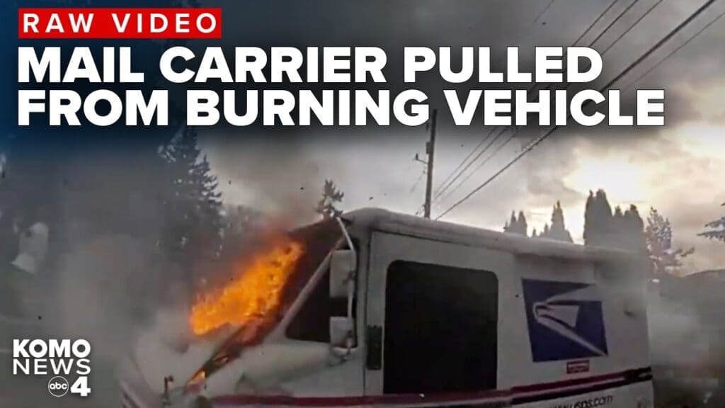 A mail delivery vehicle engulfed in flames with smoke rising in the background. The scene is set on a road with trees nearby and utility poles in the distance. Text overlay reads 'RAW VIDEO MAIL CARRIER PULLED FROM BURNING VEHICLE'.