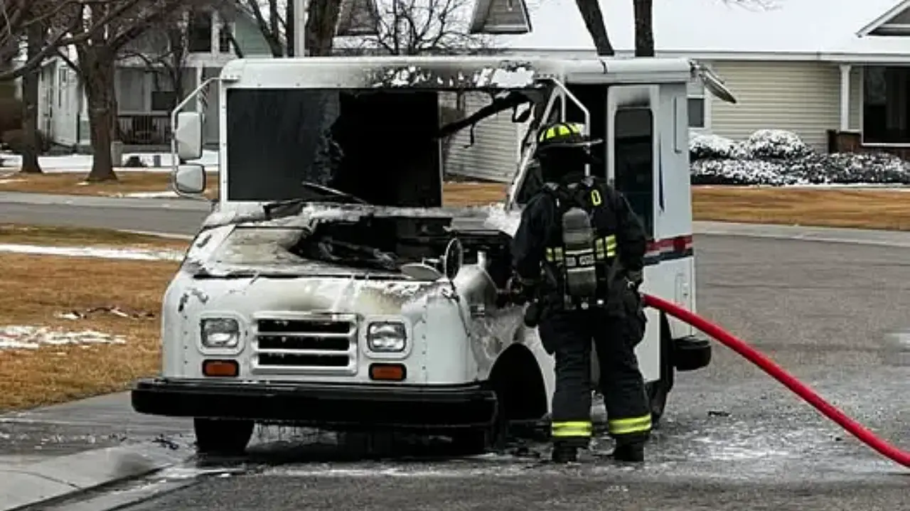 A firefighter in uniform approaches a burned postal delivery truck with smoke and damage visible on the front. Nearby, snow is on the ground and houses are in the background.