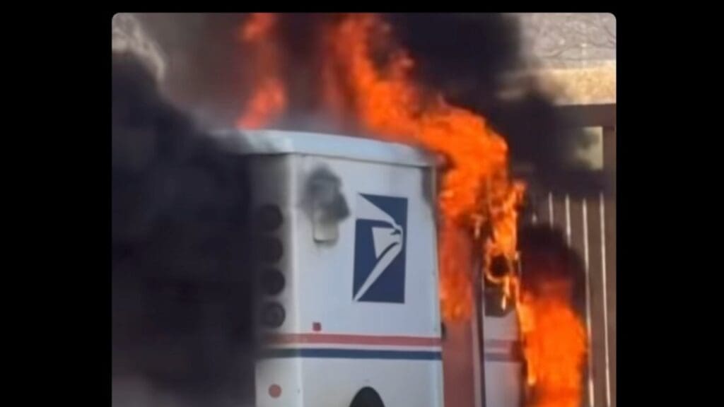 A postal service vehicle is engulfed in flames, producing thick black smoke against a blurred background. The scene depicts an incident involving the postal truck.